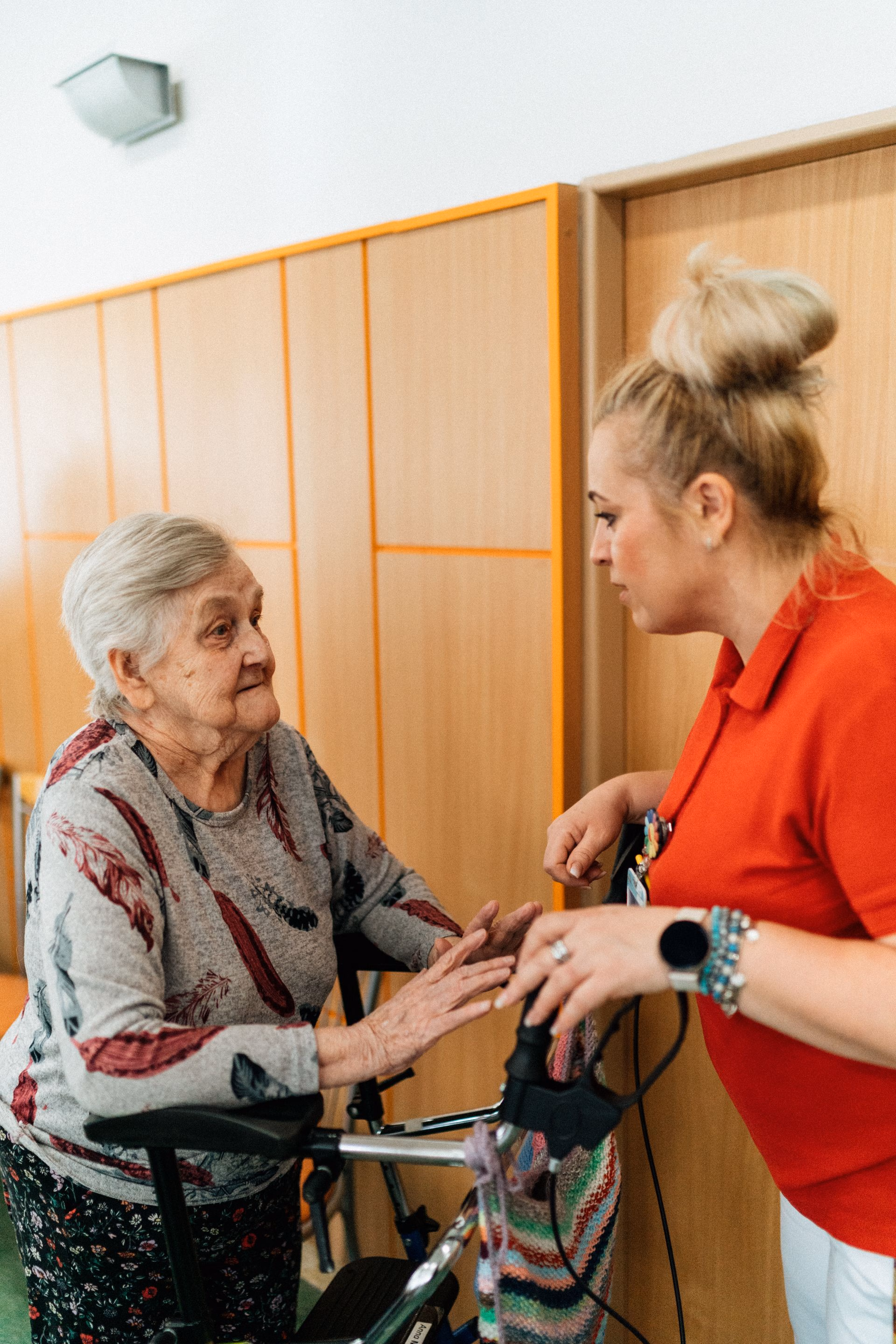 Elderly woman with walker talks to caregiver in red polo in wood-paneled room.