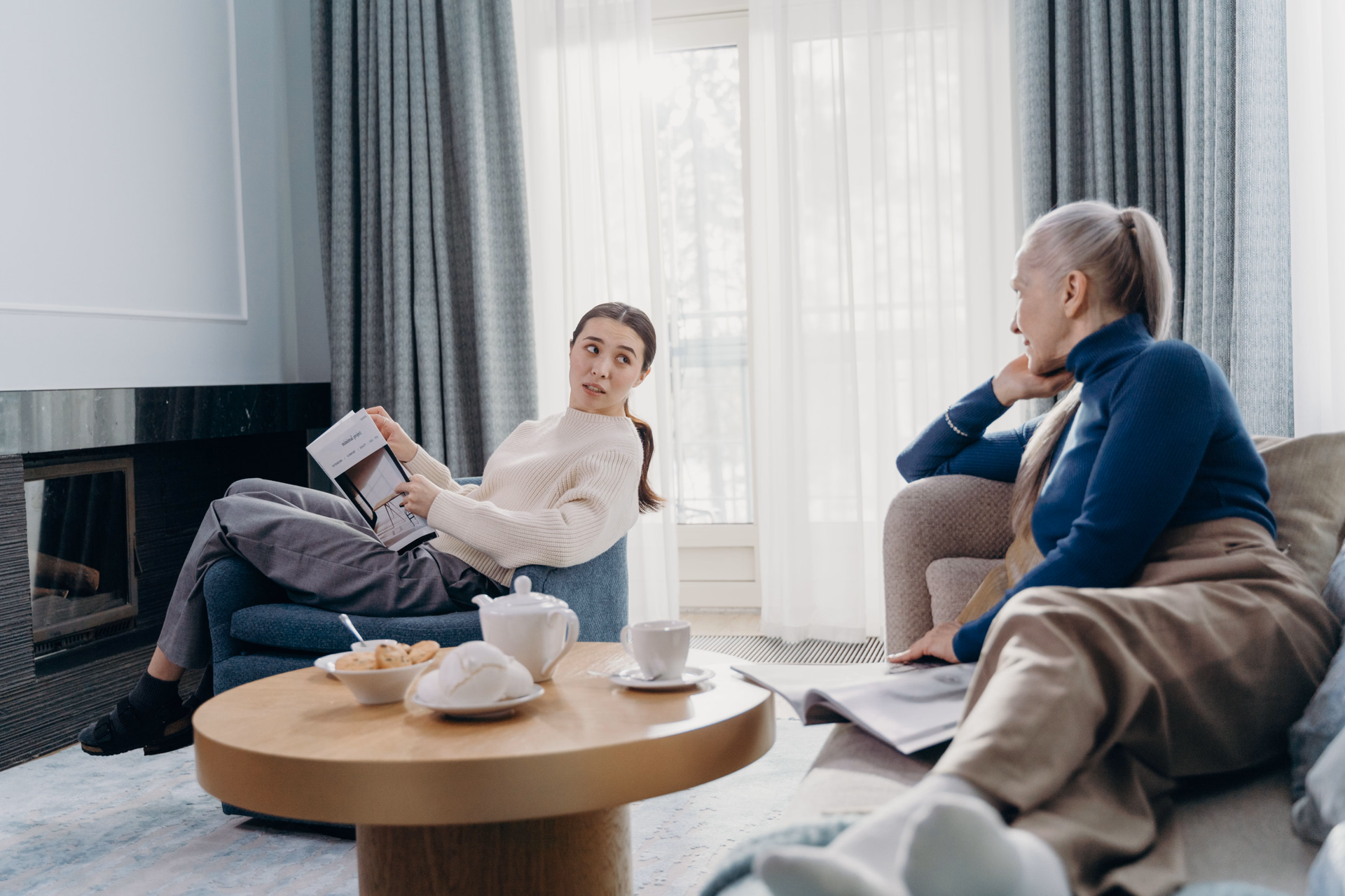 A young woman reads a magazine while an older woman relaxes on the sofa in a bright living room.