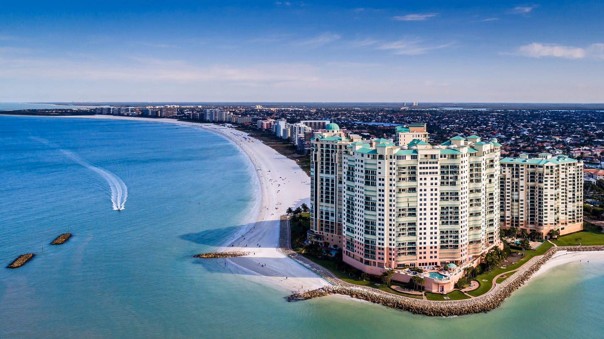 Aerial view of coastal city with beach, two boats leaving trails in water, and tall beachfront buildings.