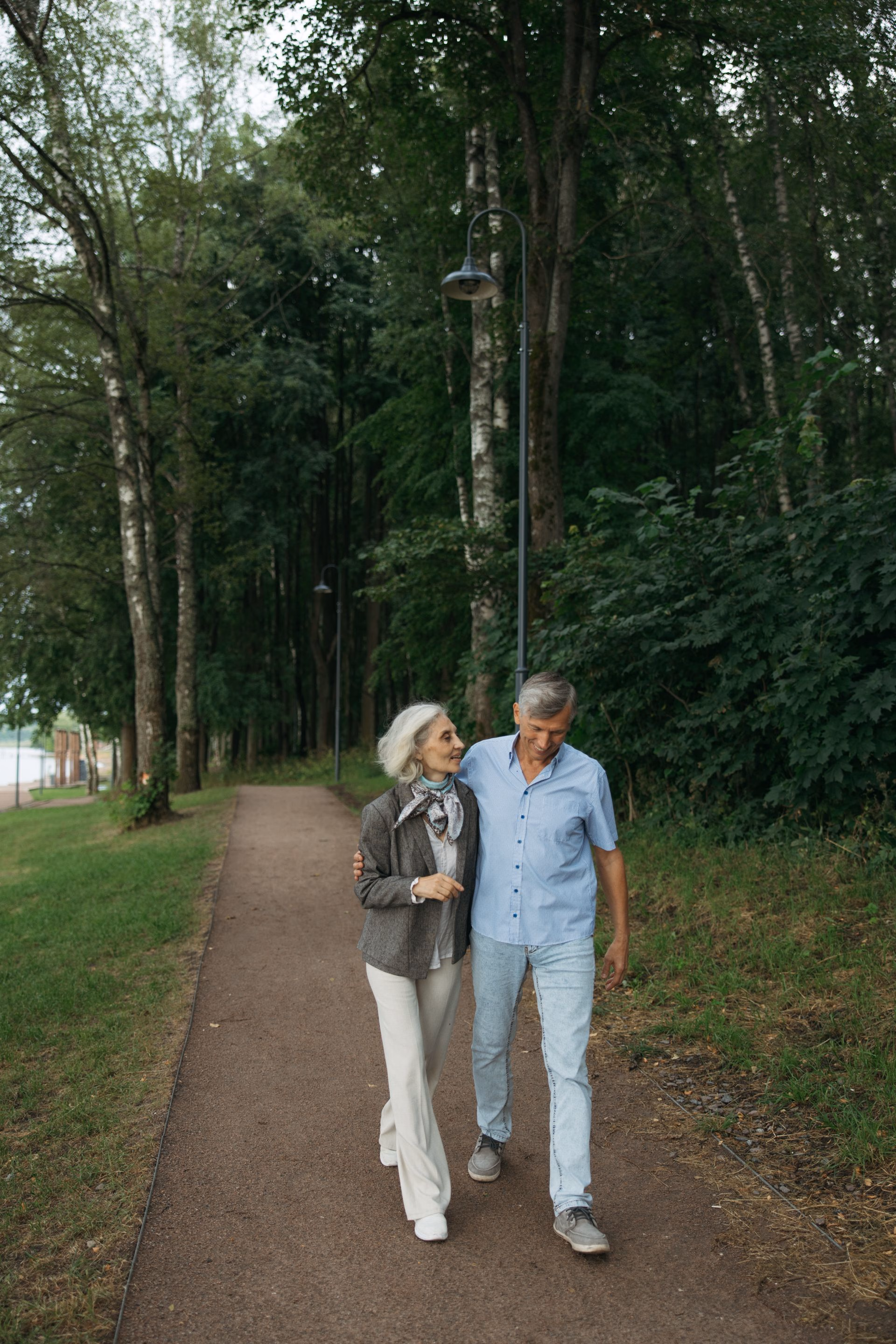 Smiling older couple walking arm in arm down a forested path with trees and grass on either side.