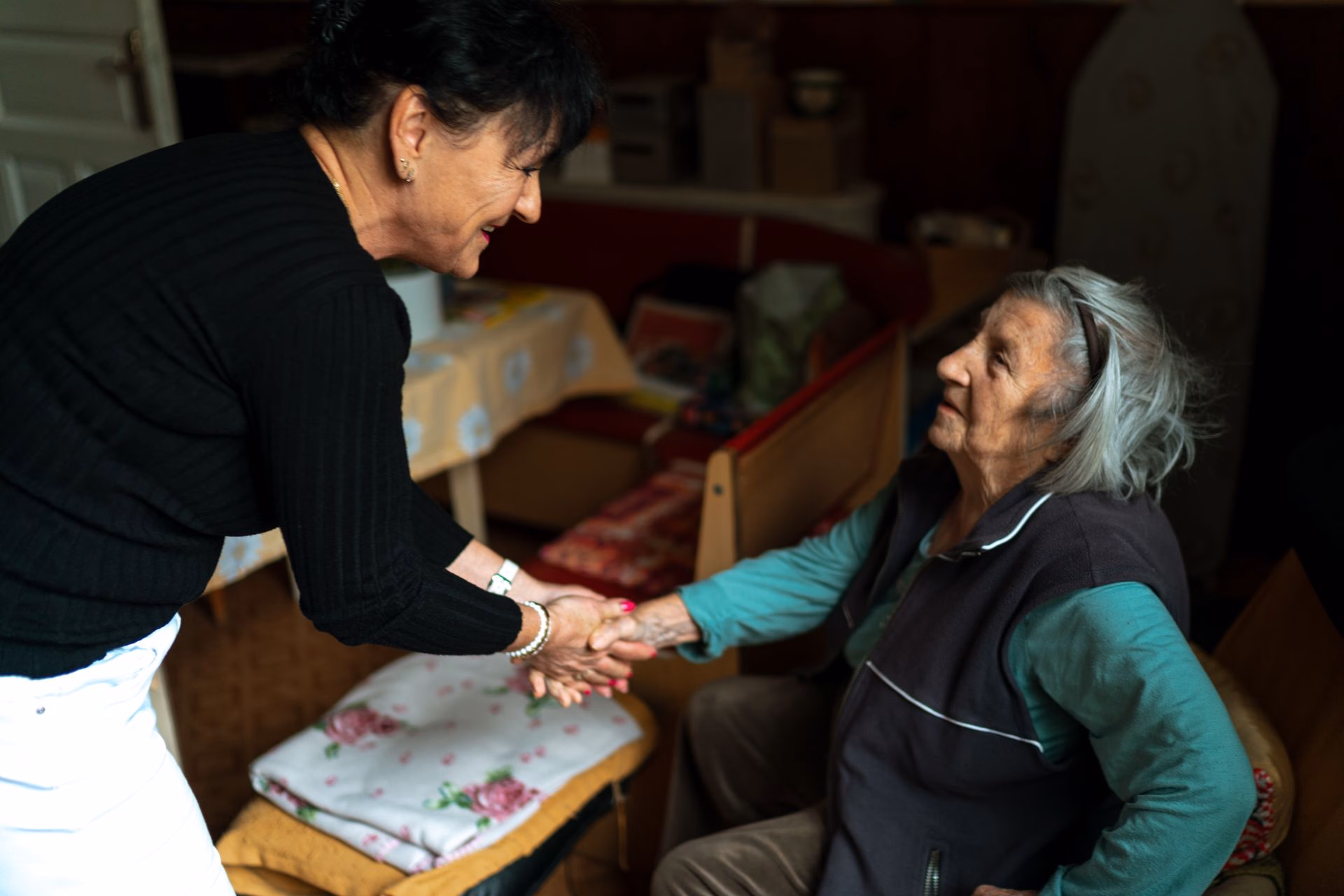 Younger woman warmly holding hands with seated elderly woman in a cozy indoor setting.