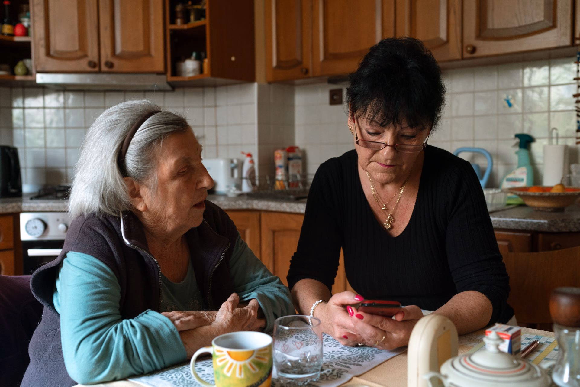 Two elderly women sitting at a kitchen table, one looking at her phone while the other watches.