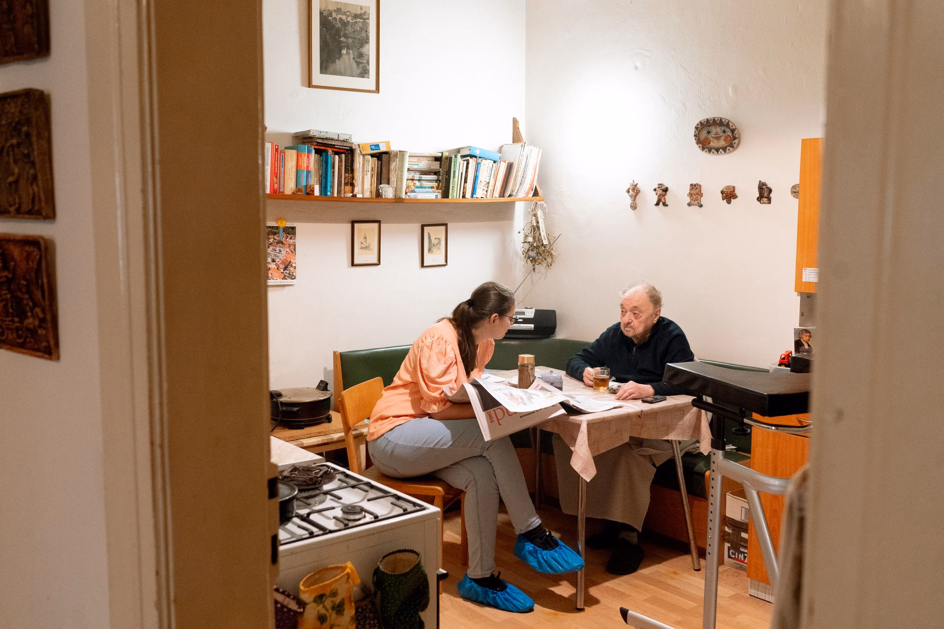 Woman talking to elderly man sitting at a kitchen table with books on a shelf behind them.