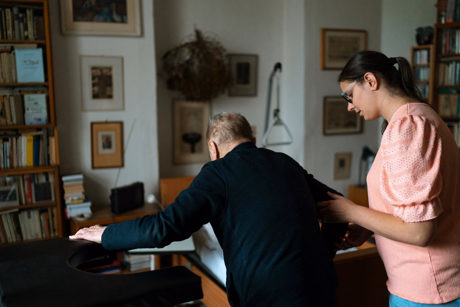 Younger woman helps elderly man stand and walk in a cozy, book-filled room at home.