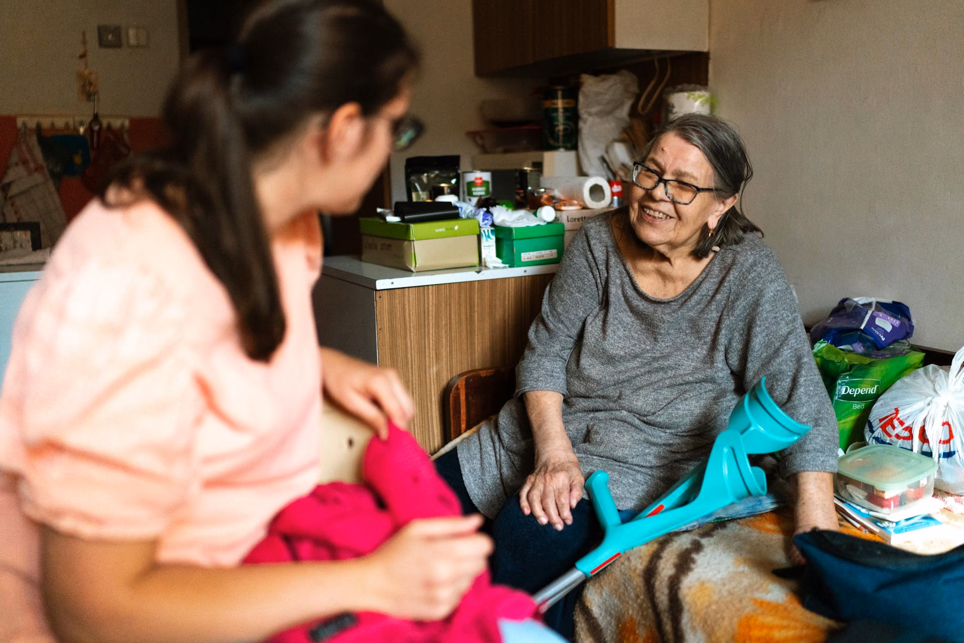Elderly woman with crutches smiling and talking to a younger woman in a cozy room.