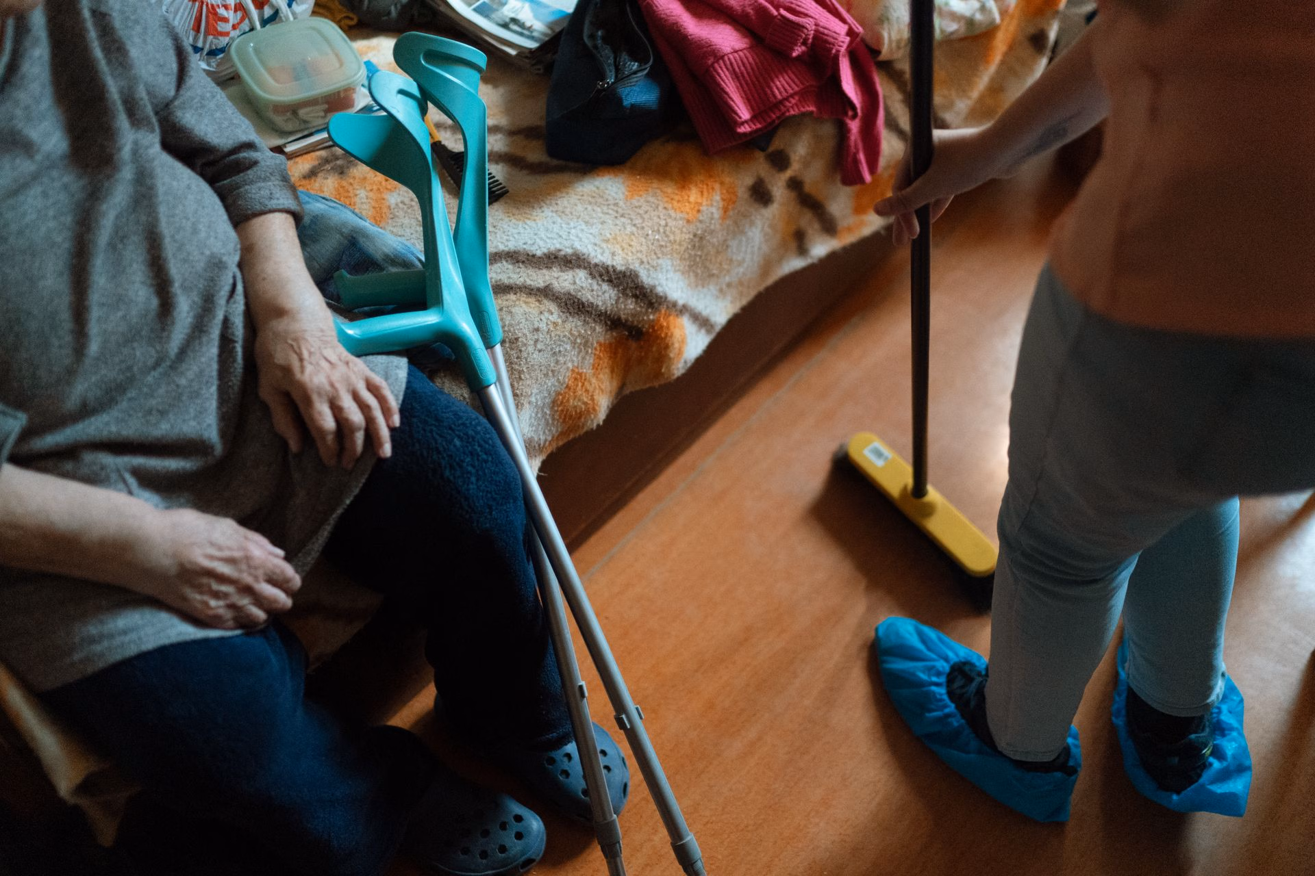 An elderly person sitting on a bed with crutches beside them and another person cleaning the floor.