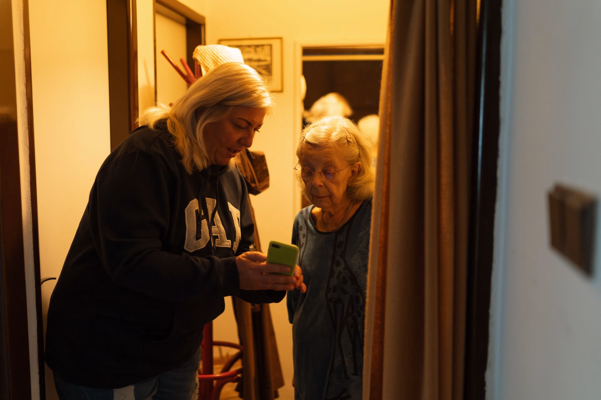 Two women standing indoors, one showing the other a phone screen, both with blonde hair.
