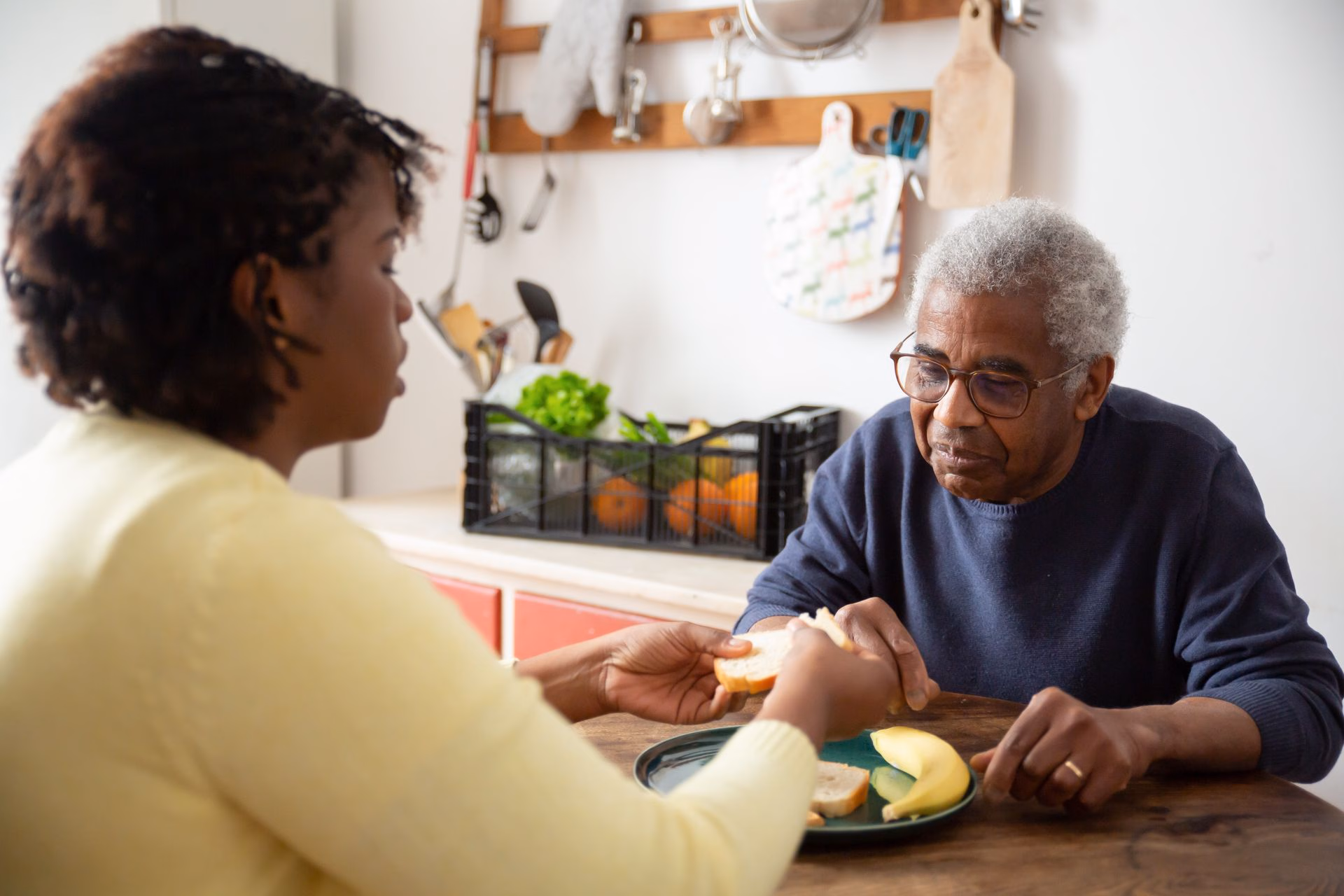 Woman handing a sandwich to an elderly man sitting at a kitchen table with a plate and banana.