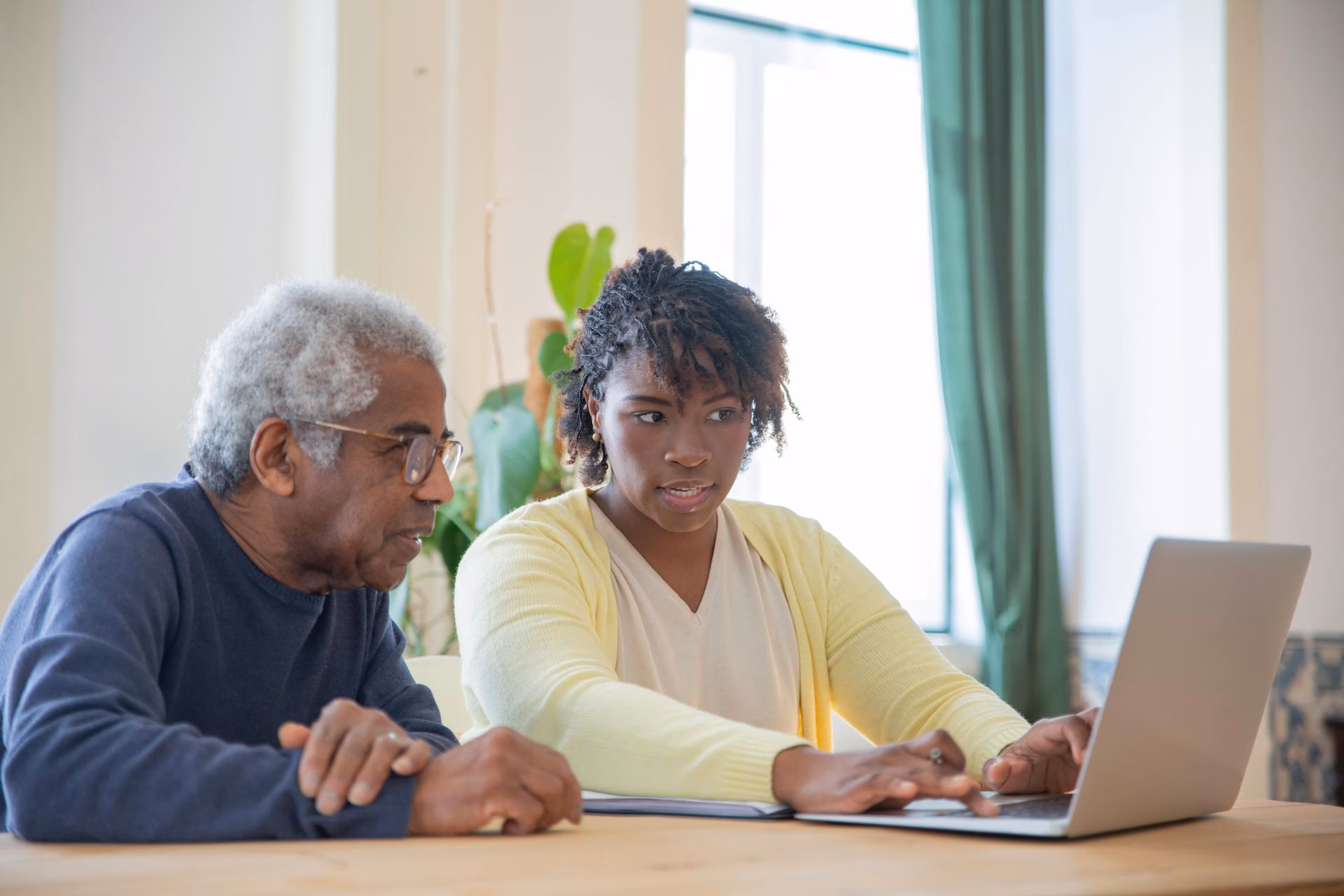 A woman showing an elderly man how to use a laptop at a wooden table in a bright room.