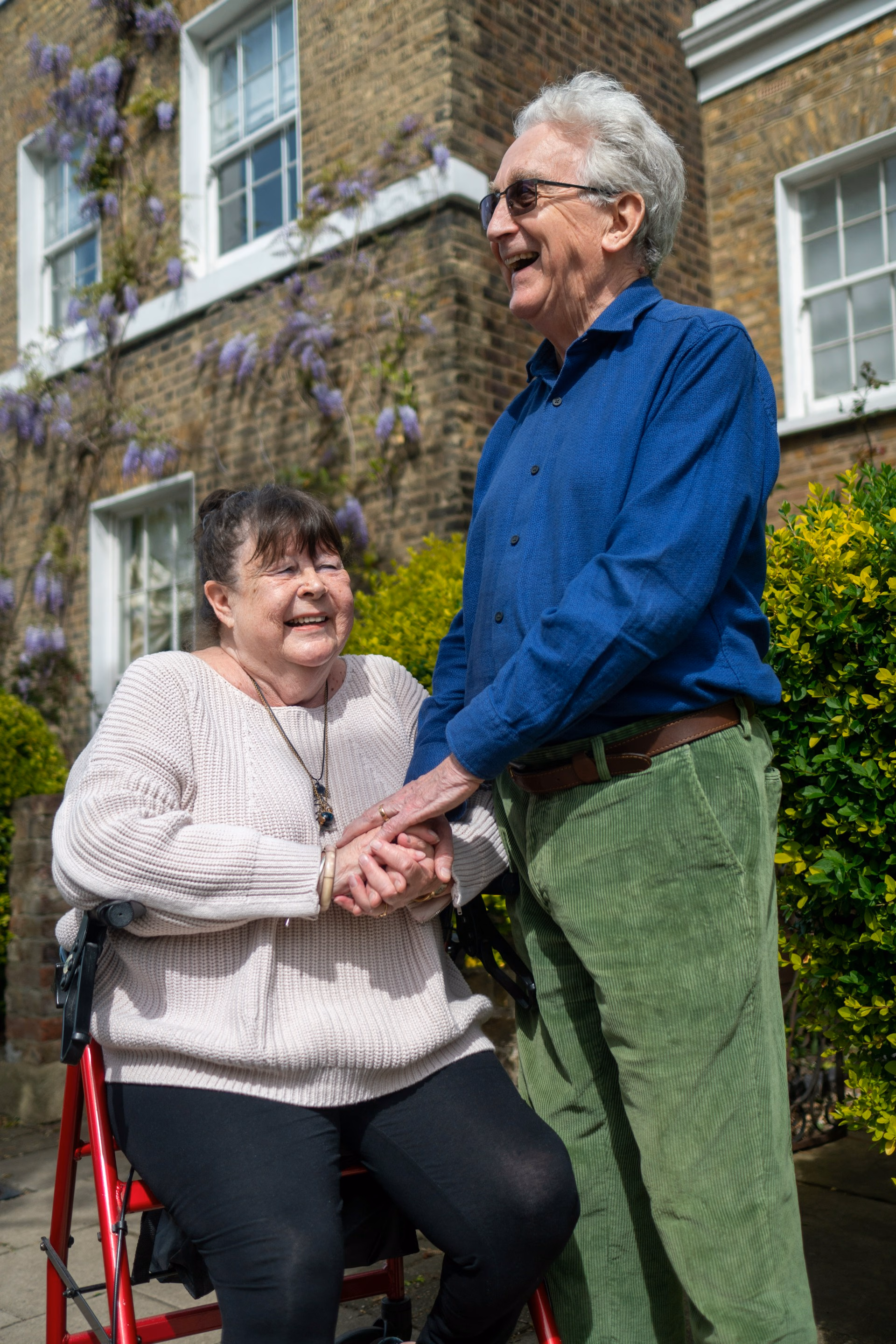 Smiling elderly couple holding hands outside a brick house with flowers and bushes.