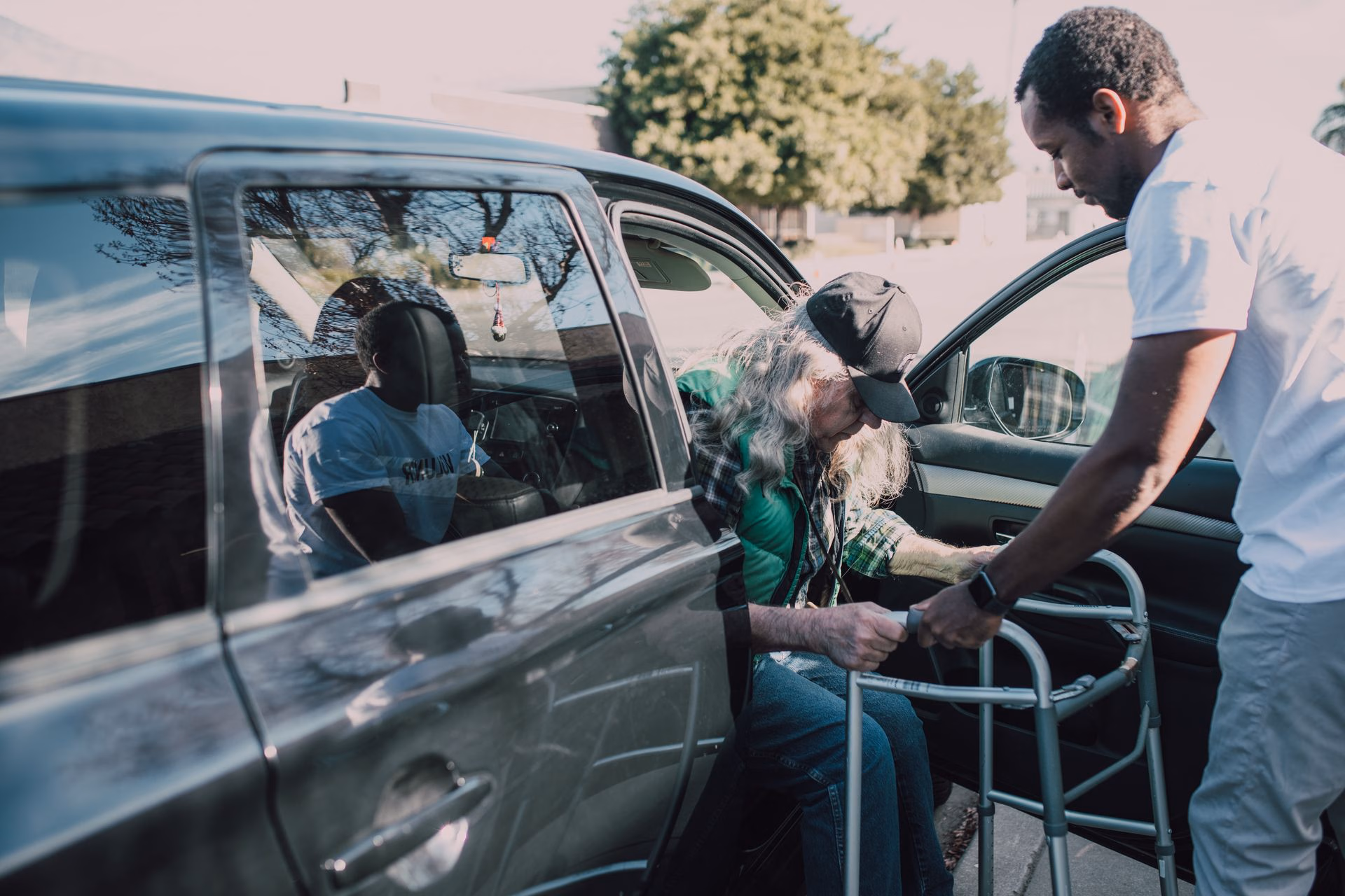 Man helps elderly woman with walker exit a parked car on a sunny day.