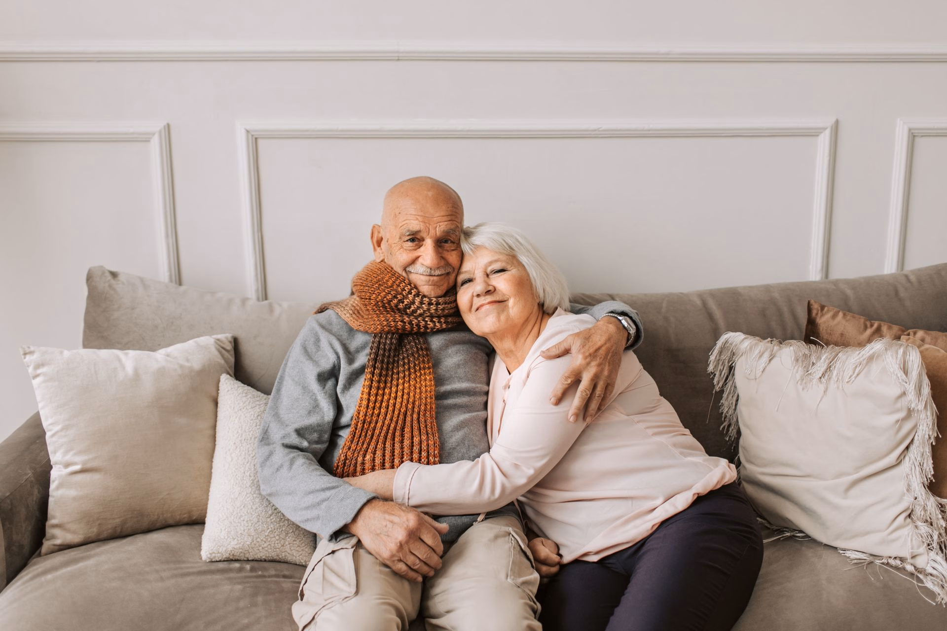 Elderly couple sitting on a couch, smiling and hugging each other warmly.