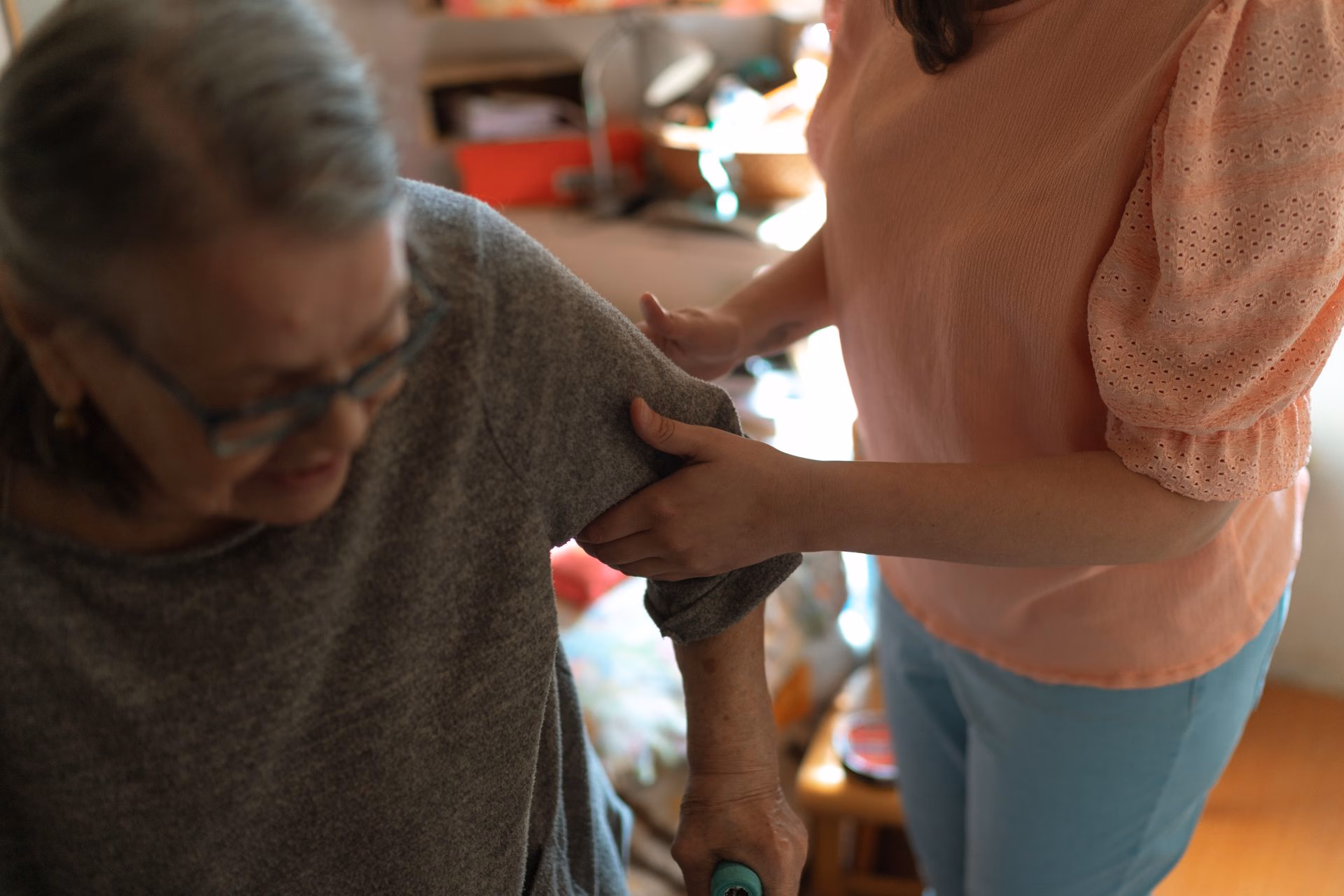 A woman in a pink blouse helps an elderly woman with gray hair and glasses to walk.
